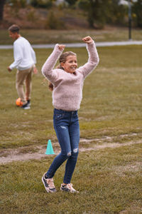 Smiling sister with arms raised standing on grassy field while brother playing soccer in background at park