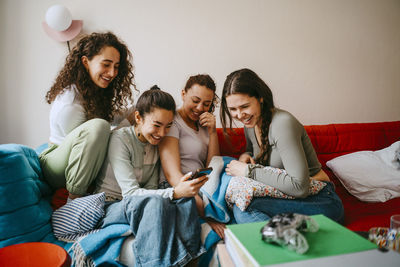 Happy young female friends looking in smart phone while sitting on sofa at home