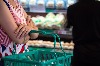 Midsection of woman holding shopping basket with man in background at supermarket