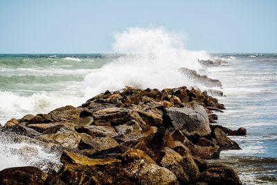 Waves splashing on rocks at shore against clear sky