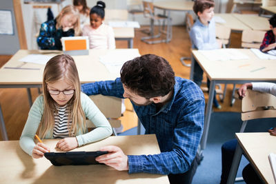 High angle view of teacher assisting female student while using digital tablet in classroom