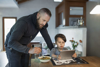 Father feeding son using digital tablet at table in house