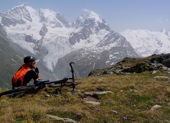 Man with bicycle sitting on hill against snowcapped mountains