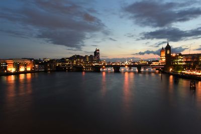 Illuminated buildings by river against cloudy sky