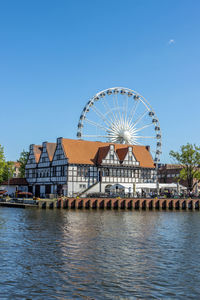Ferris wheel by the river against buildings against clear blue sky