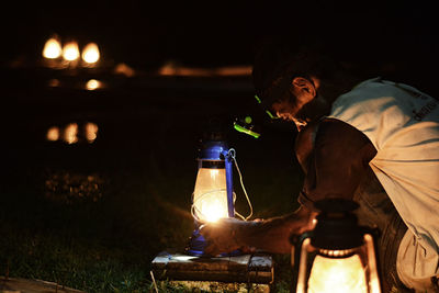 Side view of mature man with illuminated lamp while crouching on field at night