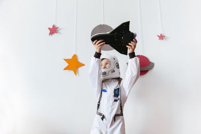 Girl wearing space suit playing with rocket against wall at home