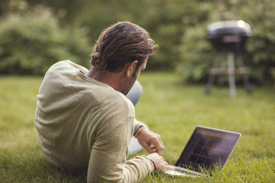 Rear view of man reclining with laptop on grass in back yard
