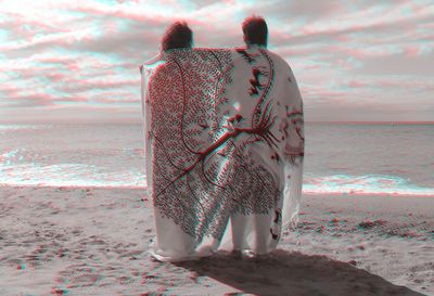 Rear view of man and woman wrapped in blanket while standing on shore at beach