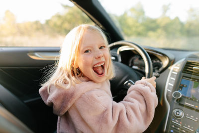 Side view of cheerful girl looking at camera sitting on driver seat of modern car with hands on steering wheel