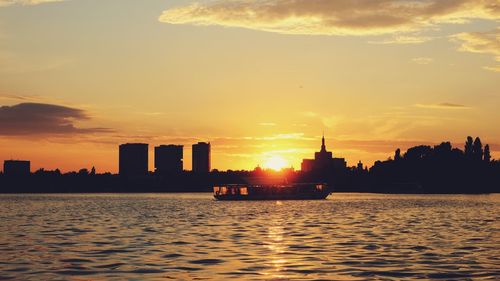 Silhouette buildings by river against sky during sunset