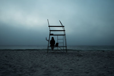 Silhouette woman sitting on lifeguard chair at beach against sky