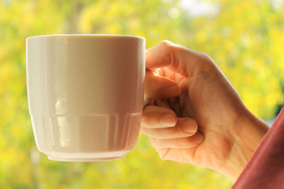 Woman's hand with a white cup close-up on a yellow-green autumn background.