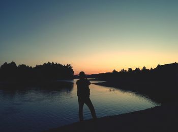 Silhouette man standing by river against clear sky during sunset