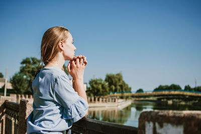 Lonely pensive young blonde woman in blue dress standing near the city river in summer sun day