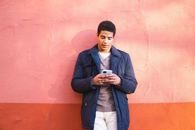 Full length of smiling man holding smart phone while standing against wall