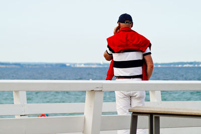 Rear view of man talking on mobile phone while standing by railing against sea