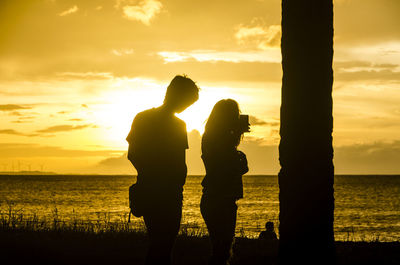 Silhouette friends standing on beach against orange sky