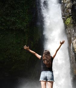 Rear view of woman with arms raised against waterfall