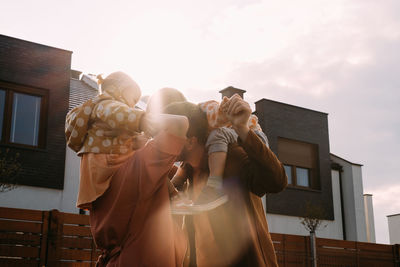 Mother father carrying children on piggyback outdoors on sunset neat house and playground. mom dad