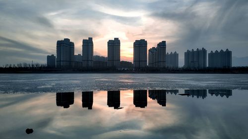 Sea by modern buildings against sky during sunset