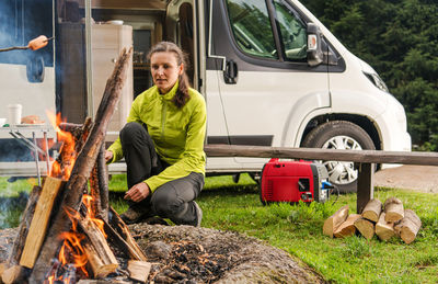 Portrait of woman crouching at campfire against motor home