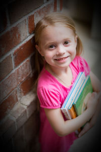 Portrait of smiling girl holding books by brick wall