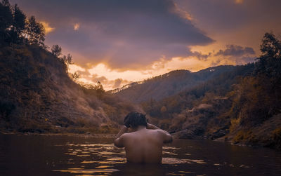 Rear view of shirtless man looking at mountains against sunset sky