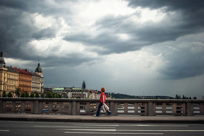 Side view of woman walking on sidewalk by railing against cloudy sky