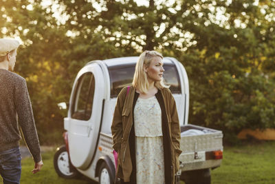 Young woman looking away while standing by man against car