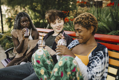 Young woman and teenage boy looking at non-binary friend while drinking in park