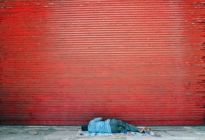 Rear view of homeless man sleeping on footpath by closed red shutter