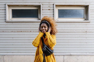 Smiling afro woman with eyes closed listening music through wireless headphones while standing against wall