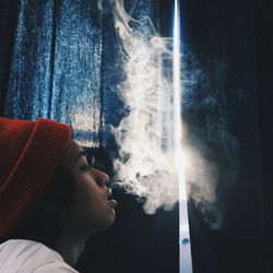 Side view of young man smoking cigarette while standing against curtain