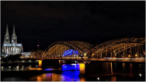 Illuminated hohenzollern bridge over rhine river by cologne cathedral against sky at night