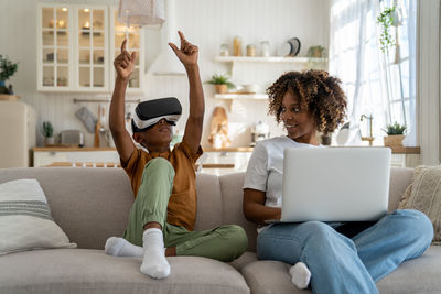 Child boy playing in virtual reality while sitting on sofa near mom working on laptop