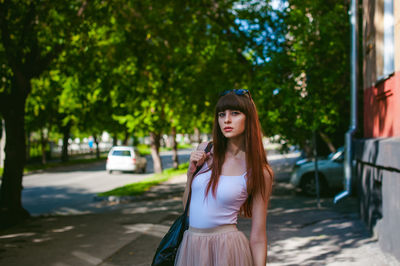 Portrait of young woman standing on sidewalk against trees