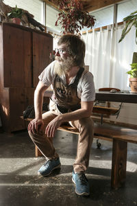 Thoughtful carpenter looking away while sitting at bench table in workshop