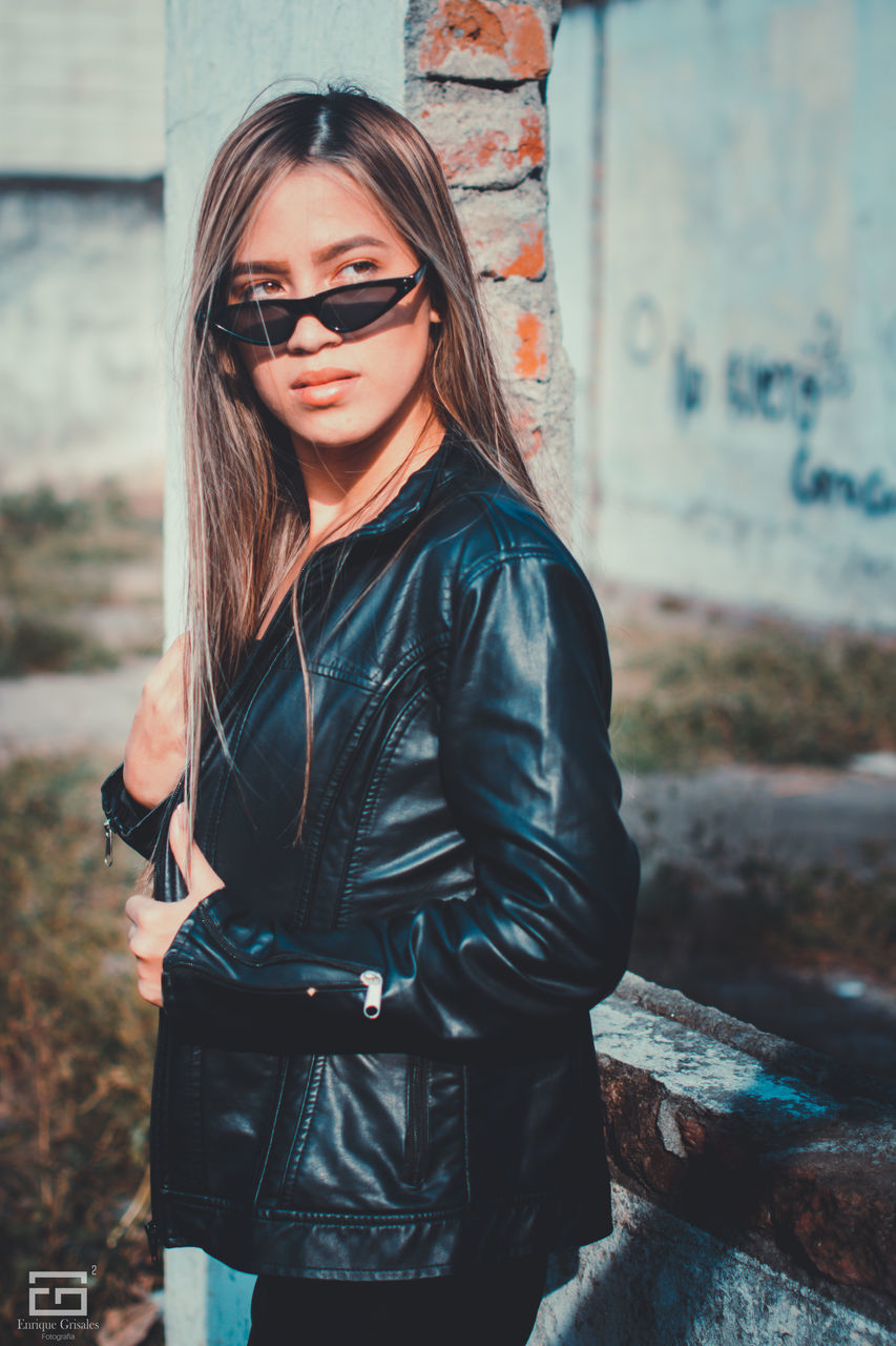 real people, one person, young adult, standing, lifestyles, young women, glasses, leisure activity, sunglasses, fashion, three quarter length, focus on foreground, casual clothing, jacket, day, women, architecture, hairstyle, leather, outdoors, beautiful woman, leather jacket