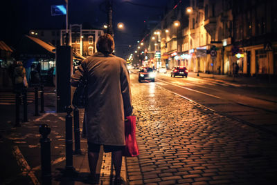 Rear view of woman standing on footpath by road in city at night
