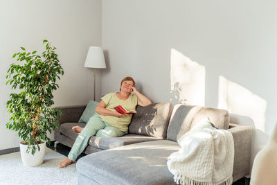 Senior plus size woman daydreaming while relaxing during reading at home on couch in living room. 