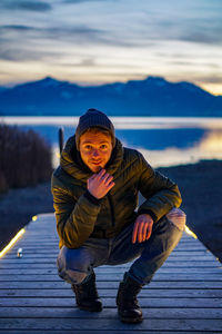 Portrait of young man crouching on pier against lake