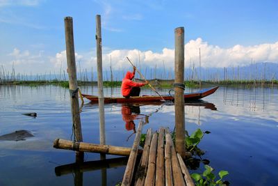 Man sitting on wooden post in lake against sky