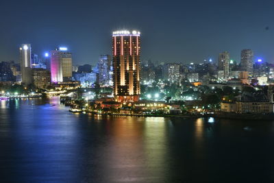 Illuminated buildings by river against sky at night