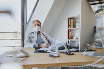 Mature doctor wearing protective face mask sitting by desk at doctor's office