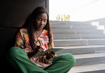 Focused young african american female student with long dark hair in casual clothes sitting on staircase in city and using tablet with stylus