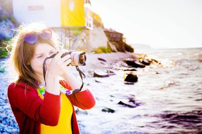 Young woman photographing with camera while standing by sea against clear sky