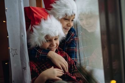 Cute children wearing snta hats and plaid shirts sitting hugging by the window waiting for christmas