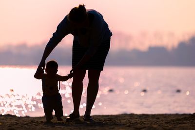 Silhouette mother holding hands of baby while standing against sea during sunset
