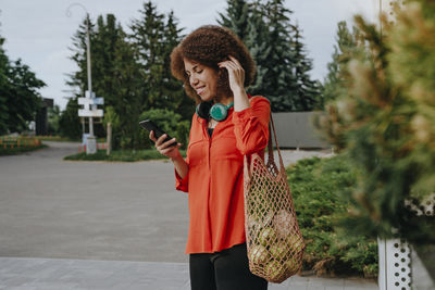 Young woman carrying apples in mesh bag and using mobile phone
